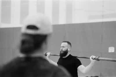 Quiet warm-up portrait of a bearded weightlifter in a black tee balancing an empty barbell across his rear deltoids in the back-rack position, eyes lowered and a single wireless earbud in place as he gathers focus before a lift, the bar held lightly in chalked hands at each shoulder, a teammate in a white cap thrown into heavy out-of-focus blur across the foreground left to frame and compress the scene, a high gymnasium wall of muted gray panels and tall windows receding softly behind, captured with shallow depth of field on Ilford HP5+ 400 black and white film whose moderate grain and rich tonal range lend the moment a contemplative, pre-competition stillness