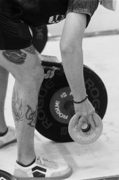 Tight, graphic warm-up detail of a heavily tattooed athlete bent over a loaded barbell, an outstretched arm with a beaded wrist band sliding a Rogue change plate onto the bar against a stack of black Rogue 25-kilogram bumper plates whose repeated white branding fans across the frame, intricate floral and octopus ink wrapping the thigh and calf, white weightlifting shoes planted on the platform below, the bleached gymnasium floor falling away into soft focus beyond, captured close on Ilford HP5+ 400 black and white film whose punchy contrast and visible grain turn the ordinary ritual of loading weight into a study of skin, steel and texture