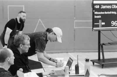 Behind-the-scenes documentary frame of the scoring and marshalling table at an Olympic weightlifting meet, a young official in a white cap leaning across the table to write attempt cards while a silver-haired coach in glasses watches intently from his seat and a shaved-headed judge sits half-cropped in the foreground, the table cluttered with a laptop, a handheld radio, a water bottle and scattered paper, the large attempt board glowing behind them with the next lifter's name and opener — James Oberle, Milwaukee Barbell, Snatch, 96 kilograms — and a singlet-and-straps athlete standing in soft focus deeper in the hall, captured on Ilford HP5+ 400 black and white film whose natural grain and balanced grays render the quiet administrative machinery that keeps a competition running