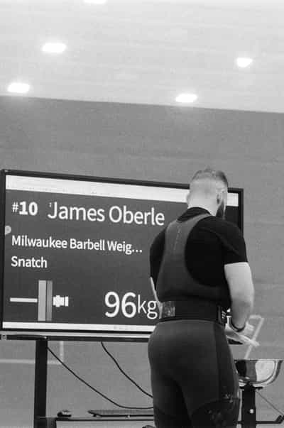 Vertical from-behind portrait of a weightlifter standing alone before the giant attempt board, his back to the camera in a black competition singlet and lifting straps, a wide leather belt cinched over chalk-dusted hips and one wrist taped, gazing up at the screen that announces his own lift — number 10, James Oberle, Milwaukee Barbell Weightlifting, Snatch, 96 kilograms — recessed ceiling lights glowing across the upper frame and a glimpse of plates and a stool at the platform's edge, captured on Ilford HP5+ 400 black and white film whose deep blacks and luminous highlights heighten the charged, solitary anticipation of a lifter measuring himself against the number waiting on the wall