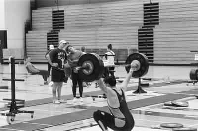 Wide environmental training-hall scene at a weightlifting meet, a lifter in the lower-right foreground sunk into a deep receiving squat with a loaded Rogue barbell held locked overhead, while coaches Nate and Jessie Stemo look on from the mid-ground — Nate in an ONYX tee and white cap with hands on hips, Jessie leaning in close beside him — among other lifters milling near the warm-up platforms, set against the long folded wooden bleachers and roll-out platforms of a school field house, plate trees and a fan scattered across the floor, captured on Ilford HP5+ 400 black and white film whose moderate grain and even gray scale capture the communal, between-attempts atmosphere of the warm-up room