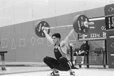 Decisive-moment competition frame of weightlifter Eric Paz in a VIRUS singlet receiving a snatch in a full overhead squat, arms locked out and the loaded barbell of black Eleiko 20-kilogram bumper plates pinned overhead in a strong diagonal across the top of the frame, face set with effort and quads driving up out of the bottom, the meet's attempt board behind him reading 121 kilograms on a second attempt beside the Fond du Lac and Cardinal venue branding, a coach or official seated in soft focus to the right, the painted field-house wall and chalked geometry sweeping behind, captured on Ilford HP5+ 400 black and white film whose grain and contrast amplify the explosive tension of the catch