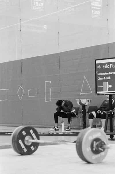 Vertical reportage frame composed through a loaded Eleiko barbell whose two great bumper-plate discs sit heavily out of focus across the foreground, drawing the eye past them to Eric Paz seated alone on a folding chair with his head bowed into his hands, gathering himself between attempts as a teammate rests beside him, the attempt board to the right glowing with number 8, Eric Paz, Milwaukee Barbell, Clean & Jerk, faded championship banners from 1922 and 1952 hanging high on the field-house wall above geometric chalk diagrams, captured on Ilford HP5+ 400 black and white film whose grain and tonal depth wrap the scene in the hushed, heavy quiet that settles over an athlete on the two-minute clock