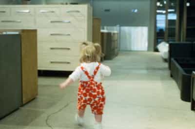 Charming toddler with blonde hair walking away through modern white kitchen wearing orange patterned overalls and white shirt, captured on CineStill 800T film with natural window lighting and contemporary cabinetry
