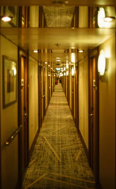 Vertigo-inducing symmetrical vanishing-point view down a seemingly infinite cruise ship passenger corridor lined with identical cabin doors and glowing circular wall sconces, patterned abstract-line carpet runway leading the eye toward a distant figure at the far end, pronounced warm amber color cast pervading the scene from daylight-balanced slide film shot entirely under tungsten lighting, overhead mirrored ceiling panels doubling the corridor's perspective and ceiling geometry, captured on Kodak Ektachrome E100 slide film with its fine grain, rich saturation, and vivid tonal separation evoking the uncanny stillness of ocean liner architecture at rest and the slightly disorienting scale of modern cruise ship interiors