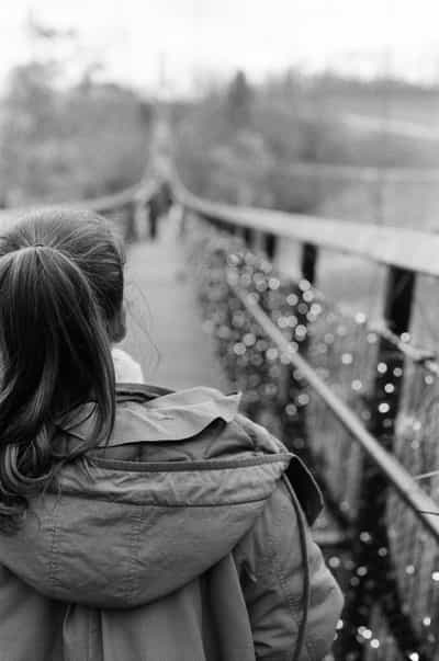 Intimate back-of-head portrait of person with long hair in ponytail wearing utility jacket gazing down wooden plank suspension footbridge, captured with extremely shallow depth of field creating dreamy circular bokeh effect on foreground bridge chains and railing, with soft-focused distant figures walking across bridge and blurred forest landscape beyond, shot on Ilford HP5+ 400 black and white film with cinematic composition and rich tonal contrast evoking contemplative solitude and childhood wonder