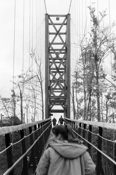 Dramatic over-the-shoulder perspective of person in hooded jacket walking toward towering suspension footbridge with geometric steel truss tower featuring industrial X-pattern support beams, wooden plank walkway with rope railings leading eye toward two distant figures, framed by tall mature trees on both sides, captured on Ilford HP5+ 400 black and white film with strong leading lines and vertical composition emphasizing architectural scale and journey symbolism