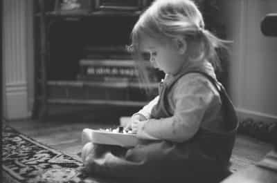 Profile view of young toddler with wispy blonde hair sitting cross-legged on hardwood floor deeply absorbed in manipulating a wooden busy board toy, captured in intimate available-light setting with upright piano softly out of focus in the background and ornate patterned area rug beneath, shallow depth of field drawing attention to the child's concentrated expression and small hands while the surrounding domestic interior falls into gentle bokeh, shot on Ilford HP5+ 400 black and white film with beautiful tonal gradation from deep rich shadows to luminous highlights, moderate film grain adding warmth and texture to this candid childhood moment of quiet discovery and independent play