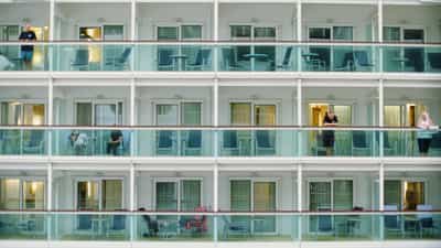 Striking architectural pattern study of cruise ship's stacked passenger cabin balconies forming a tight geometric grid across three visible tiers, teal-tinted glass railings creating rhythmic color intervals against crisp white structural dividers and identical curtained sliding doors, small candid figures occupying select balconies — a man leaning on a railing with phone in hand, a woman in black seated alone, a pair watching in shadow, a solo woman glancing out over the ocean — conveying the compressed anonymity and human scale nested within industrial hospitality architecture, repetitive arrangements of deck chairs and small tables replicated unit to unit, captured on Kodak Ektachrome E100 slide film with cool balanced palette and sharp architectural detail