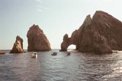 Iconic El Arco natural stone arch formation rising from turquoise Pacific waters at Land's End in Cabo San Lucas, with dramatic stratified rock formations and tourist boats navigating through the famous landmark, captured on Cyberpunk 400D film with warm vintage color tones and soft atmospheric haze creating dreamy coastal Mexico seascape