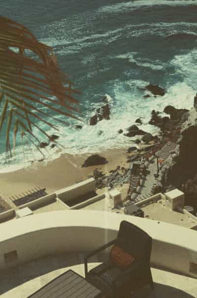 Vertiginous balcony view from luxury Cabo villa terrace overlooking dramatic rocky Pacific coastline with turquoise waves crashing against volcanic boulders, modern lounge furniture on sun-drenched patio, and wooden stairway descending to private sandy beach below, captured on FPP Retrochrome 400 film with distinctive vintage cross-processed color palette and moody blue-green tones