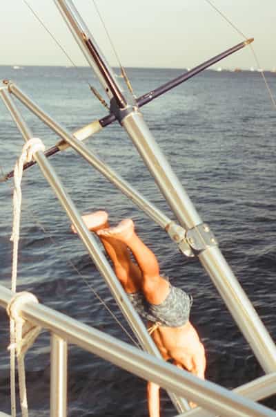 Dynamic action shot capturing person mid-dive from luxury yacht into tranquil blue-green Pacific waters off Cabo coast, with stainless steel boat rigging, white ropes, and metal railings framing the composition, shot on Cyberpunk 400D film with warm vintage tones and soft atmospheric grain creating nostalgic summer sailing adventure aesthetic