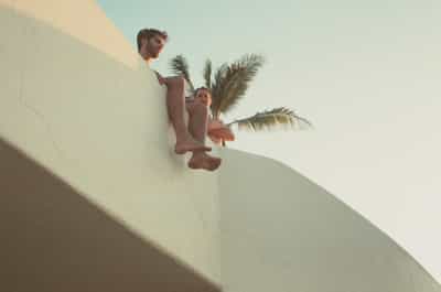 Creative upward perspective of two friends relaxing on modern white stucco villa terrace with tropical palm trees silhouetted against pale sky, geometric architectural forms framing candid vacation moment, captured on FPP Retrochrome 400 film with distinctive vintage cross-processed color palette and dreamy pastel tones creating laid-back coastal Mexico lifestyle aesthetic