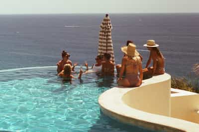 Group of friends enjoying cocktails in stunning infinity pool overlooking endless Pacific Ocean horizon, with woven sun hats, striped beach umbrella, and modern curved white architecture creating luxurious coastal villa atmosphere, captured on Cyberpunk 400D film with warm vintage tones and vibrant turquoise pool water contrasting against deep blue seascape in golden summer light