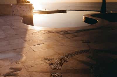 Low-angle detail shot of ornate compass rose design inlaid in weathered travertine pool deck pavers with curved white infinity pool edge and poolside furniture in background, bathed in dramatic sunrise light creating warm amber tones and deep shadows, captured on Kodak Gold 200 film with rich grain and saturated vintage color palette at luxury Cabo villa