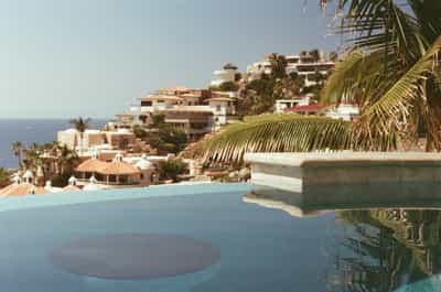 Tranquil infinity pool with pristine turquoise water and circular stone feature reflecting palm fronds, overlooking terraced Mediterranean-style villas cascading down coastal hillside toward Pacific Ocean, with thatched palapa roofs and tropical vegetation, captured on Cyberpunk 400D film with warm golden tones and soft atmospheric haze creating luxurious Baja California resort aesthetic
