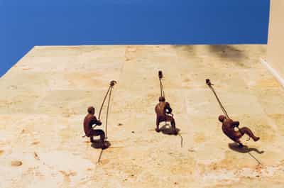 Dramatic aerial perspective of three people preparing to rappel down steep cliff face from luxury villa infinity pool edge, secured by climbing ropes against weathered travertine deck with deep blue Pacific Ocean below, harsh midday sun creating strong shadows and highlighting the adventurous contrast between resort luxury and extreme sports, captured on Kodak Gold 200 film with rich color saturation and warm coastal tones