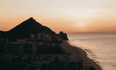 Breathtaking animated sunrise timelapse of dramatic pyramidal mountain peak silhouetted against evolving pastel sky with peach, pink, and cream gradients over Pacific Ocean, featuring Cabo resort buildings and sandy beach nestled against volcanic coastal rock formation as dawn light transforms the landscape, captured on Kodak Gold 200 film and converted to animated GIF showcasing the ethereal progression of golden hour light across Sea of Cortez coastline
