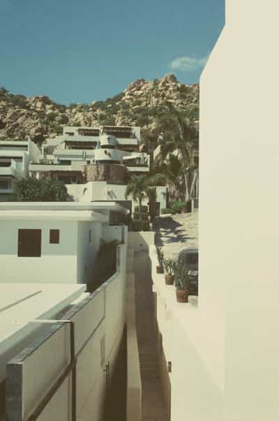 Striking architectural view of terraced white stucco villa complex with modernist design built into dramatic rocky Cabo hillside, featuring multi-level flat roofs, open terraces, tropical palm trees, and terracotta planters under bright blue sky, captured on FPP Retrochrome 400 film with vintage cross-processed aesthetic and soft pastel tones creating Mediterranean meets Baja California modern resort living
