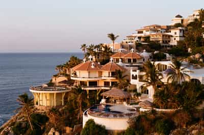 Stunning sunrise view of luxury Cabo coastal villas cascading down rocky hillside toward Pacific Ocean, featuring distinctive white stucco architecture with circular terracotta-tiled cupola buildings, thatched palapa poolside structures, modern curved terraces, and lush tropical palm trees bathed in warm golden morning light with soft pastel sky, captured on Kodak Gold 200 film with rich saturation and dreamy coastal Mexico resort aesthetic