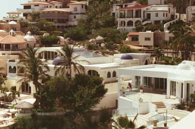 Expansive overview of luxurious Cabo resort architecture featuring terraced white stucco villas with Spanish colonial elements including domed cupolas with blue mosaic tiles, terracotta tile roofs, arched doorways, modern infinity pools, and lush tropical palm trees nestled into coastal hillside, captured on Cyberpunk 400D film with warm golden tones creating idyllic Baja California Mediterranean-style vacation paradise