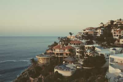 Breathtaking sunrise view of terraced Cabo coastal villas with distinctive circular terracotta-roofed buildings, white stucco architecture with Spanish colonial elements, tropical palm trees, and dramatic rocky coastline meeting tranquil Sea of Cortez under soft pastel dawn sky, captured on FPP Retrochrome 400 film with ethereal vintage cross-processed aesthetic and warm golden light creating serene Baja California morning paradise