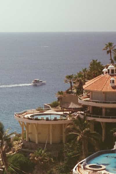 Luxury motor yacht cruising through calm blue Pacific waters past terraced Cabo coastal villas featuring Spanish colonial architecture with white domed cupolas, terracotta tile roofs, curved infinity pools, and lush tropical palm trees cascading down rocky hillside, captured on Cyberpunk 400D film with soft atmospheric grain and vintage warm tones creating quintessential Baja California luxury lifestyle aesthetic