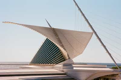 Three-quarter view of the Milwaukee Art Museum's Quadracci Pavilion seen from the lakefront terrace, Santiago Calatrava's brise soleil wing rising and curling overhead in a dramatic sweep of white louvered fins while the angled cable-stayed mast of the pedestrian bridge slices diagonally across a clear pale sky, the building's glass-walled reception hall glowing dark teal beneath the structure and the flat steel-blue line of Lake Michigan stretching across the horizon behind, low foreground rooflines and walkways leading the eye up into the soaring form, captured on Kodak Ektar 100 whose vivid saturation and razor-fine grain sharpen the interplay of taut cables, curved ribs and open water into a study of motion frozen in architecture.