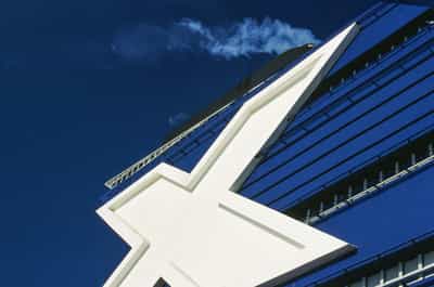 Bold low-angle graphic study of the towering white Celebrity Cruises chi emblem — the Greek letter X rendered as two crisp intersecting beveled strokes with hollow inner outlines — mounted high on the deep cobalt-blue hull of a moored cruise ship, the angular geometry of the mark carving sharp negative space against the saturated navy paneling and rhythmic horizontal cabin window slits receding toward the right edge of the frame, a single wisp of high cirrus cloud drifting through the richly saturated tropical sky in the upper left, faint contrail trace barely visible at the lower-left horizon, shadow of the ship's funnel just clipping the topmost arm of the chi and grounding the brand mark in the architecture of the vessel itself, captured on Kodak Ektachrome E100 slide film with vivid color separation between the chalk-white emblem and the deeply saturated navy hull conveying the corporate iconography of cruise line branding rendered with the patience and richness of analog film