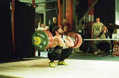 Intense action shot of bearded athlete in deep squat position catching heavy barbell with colorful Eleiko weight plates during Olympic weightlifting competition, wearing bright neon yellow lifting shoes, surrounded by spectators and judges in garage-style CrossFit gym with exposed beams and natural lighting, captured on Kodak Portra 800 film with warm vintage tones and rich grain emphasizing raw athleticism and competitive energy