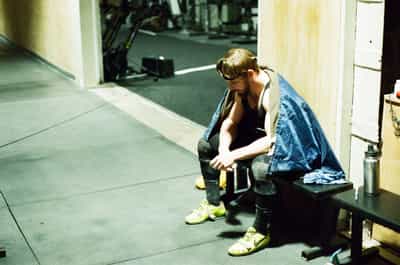 Quiet candid portrait of a bearded competitive Olympic weightlifter in black tank top and lifting belt seated on a utility bench in a gym pathway, head bowed in solitary concentration between attempts, blue towel draped over his shoulder like a cape, bright chartreuse Nike Romaleo lifting shoes grounding the composition against the dark rubber-matted platform floor, stainless steel water bottle and discarded hoodie on the bench beside him, competition rack and loaded barbells visible in soft focus through the doorway behind, warm cream gym walls catching harsh overhead light, expansive empty floor surrounding the lifter emphasizing his isolation, captured on Portra 800 film with fine grain and natural skin-tone rendering evoking the introspective stillness of competition downtime and the weight of mental preparation before a lift