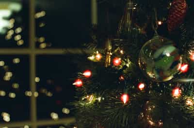 Intimate close-up of illuminated Christmas tree featuring glowing red vintage candle-style lights and green glass ornament with rich bokeh from window lights in background, fresh evergreen pine needles and holiday decorations creating warm nostalgic atmosphere, captured on Kodak Ektachrome E100 slide film with saturated vintage color palette and distinctive film grain, evoking traditional holiday magic and cozy winter evening ambiance
