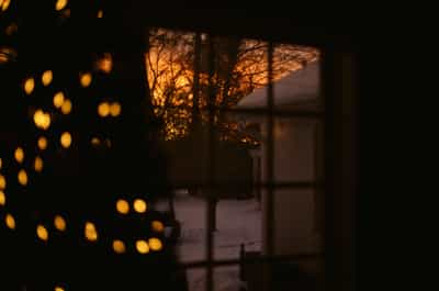 Atmospheric interior view through multi-pane window capturing dramatic golden hour sunset sky with silhouetted bare winter trees, Christmas tree lights creating dreamy golden bokeh orbs in left foreground, combining cozy holiday interior warmth with moody winter landscape beyond the glass, captured on Kodak Ektachrome E100 slide film with rich amber and deep black tonal contrast, evoking contemplative holiday evening solitude and the magic of winter twilight