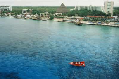 Elevated perspective from cruise ship deck looking down at a lone red passenger speedboat skimming across deep navy and turquoise Cozumel waters toward a sprawling coastal resort complex, a distinctive pyramidal thatched-roof central pavilion anchoring the palm-lined shoreline flanked by terracotta-roofed villas and a modern multi-story beachfront hotel tower, dark tropical vegetation forming a lush ridge behind the development, overcast sky softening the light across the entire scene and flattening tonal contrast, small white launch boats dotted along the resort's private pier, captured on Kodak Ektachrome E100 slide film with rich water saturation and compressed highlights conveying the aerial intimacy of approaching a Caribbean port of call