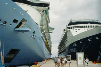 Dramatic street-level perspective of two massive cruise ship hulls flanking a narrow dockside walkway — the navy-blue Freedom of the Seas looming on the left with its fortress-like superstructure and tiered lifeboats, and the dark indigo Celebrity Summit on the right with its characteristic white crown bridge — small groups of passengers in summer clothes wandering between the vessels toward the photographer, bright blue mooring ropes and traffic-orange bollards punctuating the bleached concrete pier, overcast Caribbean sky and a narrow sliver of ocean visible in the wedge between ships, scale contrast between human figures and towering steel reinforcing the industrial immensity of modern cruise tourism, captured on Kodak Ektachrome E100 slide film with natural saturation and sharp architectural detail