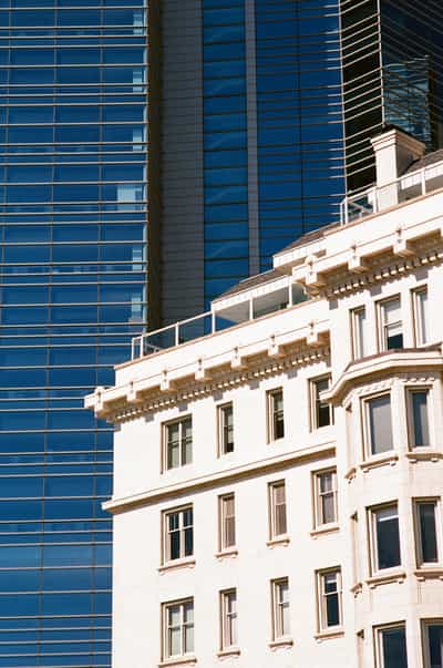 Tight architectural juxtaposition in downtown Milwaukee pairing old and new, the cream terra-cotta corner of the historic Cudahy Tower — its Beaux-Arts cornice, balustraded setback and regularly punched windows catching warm sunlight — set hard against the deep cobalt glass-and-steel curtain wall of the modern Northwestern Mutual tower rising behind it, the reflective mullioned facade receding in cool blue verticals that throw the older building's pale ornamented masonry into crisp relief, the frame cropped so the two eras of construction press directly against one another, captured on Kodak Ektar 100 whose fine grain and rich saturation deepen the electric blue glass and render the creamy stonework with clean, sunlit clarity.