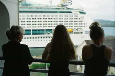 Tender back-of-head triple portrait of three women in matching black tops arranged in loose symmetry along a white balcony railing — on the left a woman with graying hair pulled back and gold hoop earrings in a cropped blazer, in the center a woman with long cascading blonde hair down her back, on the right a woman with a high blonde topknot in a ribbed tank — all gazing together toward the massive white Royal Caribbean cruise ship docked across the channel in the mid-distance, rolling green Caribbean hills rising beyond, natural overcast side light gently modeling hair and shoulders without harsh shadow, captured on Kodak Ektachrome E100 slide film with muted natural palette and shallow cinematic focus evoking shared family presence and collective anticipation of departure