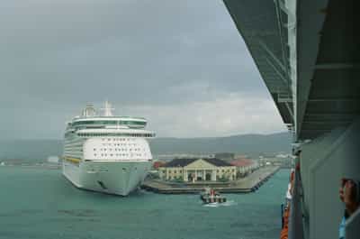 Brooding overcast arrival at Falmouth Jamaica's cruise port captured from the starboard deck of an approaching ship, the white hull and prominent navigation bridge of a docked Royal Caribbean vessel anchoring the middle distance, a small tugboat with an orange superstructure churning through choppy blue-gray harbor waters in the foreground, the pale yellow classical-columned port terminal building catching what little light breaks through the dense cloud cover, misty green Jamaican mountain ridges receding into atmospheric haze behind the port, tourist hands and a camera lens barely visible in the right foreground conveying the shared voyeuristic act of arrival, captured on Kodak Ektachrome E100 slide film with moody muted palette and flattened tonal range evoking working-day documentary travel