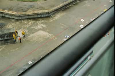 Strong geometric overhead documentary composition of three Jamaican dockhands in fluorescent yellow safety jackets and hard hats gathered in conversation near the bow of a cruise ship, their small figures dwarfed by the weathered concrete expanse of Falmouth pier with its hand-painted traffic-red safety line slicing a vivid diagonal across the frame, faded stenciled ID markers and block numerals scattered across the pavement reading as abstract signage, the heavy black painted steel railing of the photographer's ship cutting a dominant diagonal through the lower-right third of the frame and creating layered architectural perspective, muted industrial palette of concrete grays, safety yellows, and utility reds captured on Kodak Ektachrome E100 slide film with sharp detail evoking classic documentary photography of port labor