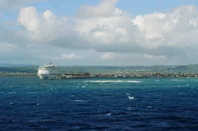 Wide panoramic view of the Royal Caribbean cruise ship Freedom of the Seas docked at Falmouth Jamaica's terracotta-roofed port complex, seen from open water with the deep blue Caribbean Sea rippling with scattered whitecaps in the foreground and the emerald-green Jamaican mountain ridges receding into the horizon, broken cumulus clouds drifting across a soft blue sky with diffused late-morning light, small navigation buoys and channel markers punctuating the turquoise-to-sapphire gradient of the water, the bright white ship anchoring the composition against the layered tropical landscape, captured on Kodak Ektachrome E100 slide film with rich water and sky saturation evoking classic travel photography of Caribbean ports of call