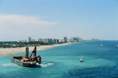Sweeping aerial perspective of Fort Lauderdale's coastline from cruise ship altitude, with a large red-and-black industrial dredging barge churning through turquoise shallows in the lower foreground, the long golden expanse of beach receding into the distance dotted with tiny beachgoers beneath colorful umbrellas, a dense wall of pastel high-rise resort towers and condominiums forming the characteristic Florida skyline behind, scattered sailboats and a small motor yacht dotting the deeper blue offshore waters, tropical palms lining the beach edge, crisp midday Florida sunlight rendering everything in bright primary colors, captured on Kodak Ektachrome E100 slide film with saturated daylight palette evoking the iconic gateway view of departing South Florida by sea