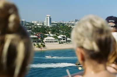 Intimate over-the-shoulder composition with two out-of-focus family members' heads framing the left and right foreground as a sharply focused small powerboat speeds across the calm aqua waters of the Fort Lauderdale inlet toward palm-lined private waterfront mansions on the sandy shore opposite, the distinctive white-capped cylindrical Pier 66 tower rising from the cluster of pastel high-rises in the middle distance, bright Florida sun illuminating the entire scene with crisp primary colors, shallow depth of field emphasizing the layered relationship between the observers in foreground and the scene they share, captured on Kodak Ektachrome E100 slide film with warm daylight palette conveying shared vacation spectatorship and departure anticipation