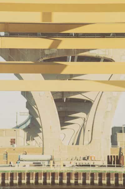Abstract upward study of the underside of Milwaukee's Hoan Bridge, the tied-arch viaduct's pale concrete deck girders stacking across the frame as broad horizontal bands while the curving ribs and box beams of its substructure sweep and converge in the gaps between them, sunlight raking the soffits into planes of cream and shadow, a glimpse of riverfront industry — a brick building, tanks and a steel sheet-pile bulkhead along the water — anchoring the base of the composition, the layered geometry flattened into a near-graphic rhythm of stripes and arcs, rendered on cross-processed FPP Retrochrome 400 slide film whose yellow-shifted, faded palette and soft grain turn the massive infrastructure into a warm, dreamlike abstraction.