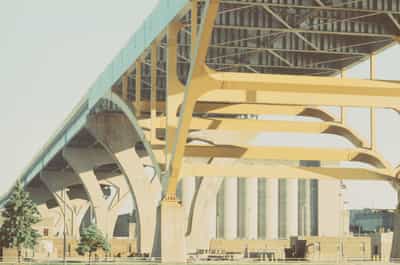 Low-angle perspective gazing up the flank of Milwaukee's Hoan Bridge, the tied-arch span receding in steep diagonal recession with its pale blue deck edge and golden-yellow steel cross-bracing marching toward a distant vanishing point, slender concrete piers fanning out beneath the roadway and a cluster of cylindrical grain elevators and low industrial buildings filling the Inner Harbor flats below, a few evergreen trees and a guardrail grounding the foreground, all beneath a washed pale sky, captured on cross-processed FPP Retrochrome 400 whose desaturated, vintage color shift and fine grain lend the muscular freeway structure a quiet, sun-faded grandeur and emphasize the rhythmic repetition of its supporting bents.