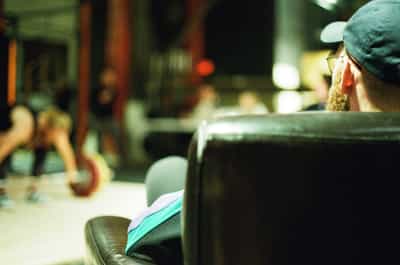 Intimate over-the-shoulder portrait of competition judge wearing green cap and teal jacket observing Olympic weightlifting meet, shot with shallow depth of field creating dreamy bokeh of blurred athletes and garage gym equipment in background, captured on Kodak Portra 800 film with warm muted tones and soft grain, emphasizing the documentary perspective and contemplative atmosphere of competitive weightlifting judging