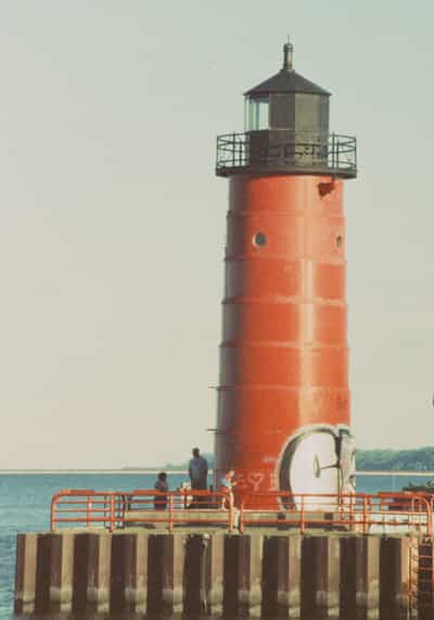 Vertical portrait of Milwaukee's historic Pierhead Lighthouse rising from its breakwater, the cylindrical red-painted tower with riveted steel plates, a circular porthole and a black railed gallery beneath its lantern dome dominating the right of the frame, a small cluster of visitors gathered at its base along a red pipe railing atop the timber-and-stone pier, faded graffiti scrawled across the tower's lower flank, and the calm steel-blue expanse of Lake Michigan opening to a low tree-lined far shore on the left, captured on cross-processed FPP Retrochrome 400 whose muted pastel tones, soft contrast and vintage color cast wrap the lakefront landmark in a warm, time-worn nostalgia.