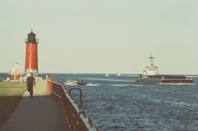 Documentary harbor scene along a Milwaukee breakwater pier, a lone man in dark clothing walking toward the camera down the concrete walkway beside a red pipe railing while the candy-red Pierhead Lighthouse with its black lantern room stands sentinel on the left, a small open motorboat carving a wake through the channel and a working tugboat shoving a long black barge across the right half of the frame, distant sailboats and a breakwater beacon dotting the seam where Lake Michigan meets a hazy sky, the whole scene rendered in the cool, faded, slightly off-register palette of cross-processed FPP Retrochrome 400 slide film whose vintage color shift and gentle grain lend the everyday waterfront a nostalgic, sun-bleached stillness.