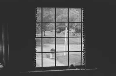 Tender black and white interior photograph of young toddler peering over window sill through traditional multi-pane window with decorative curtains, gazing contemplatively at pristine summer lawn with ornamental white pillar, manicured gardens, and mature trees beyond, capturing quiet childhood moment of wonder and discovery in cozy home interior, shot on Ilford HP5+ 400 film with rich tonal contrast and timeless monochrome aesthetic