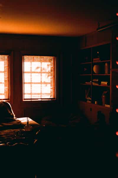 Moody atmospheric interior featuring warm amber window light illuminating built-in bookshelves, reading chair, and scattered papers in contemplative home office setting, photographed on Aerocolor IV film