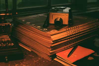 Nostalgic desk scene featuring vintage orange camera resting on stack of Fortune magazines with warm ambient lighting creating rich shadows and textures, photographed on Aerocolor IV film